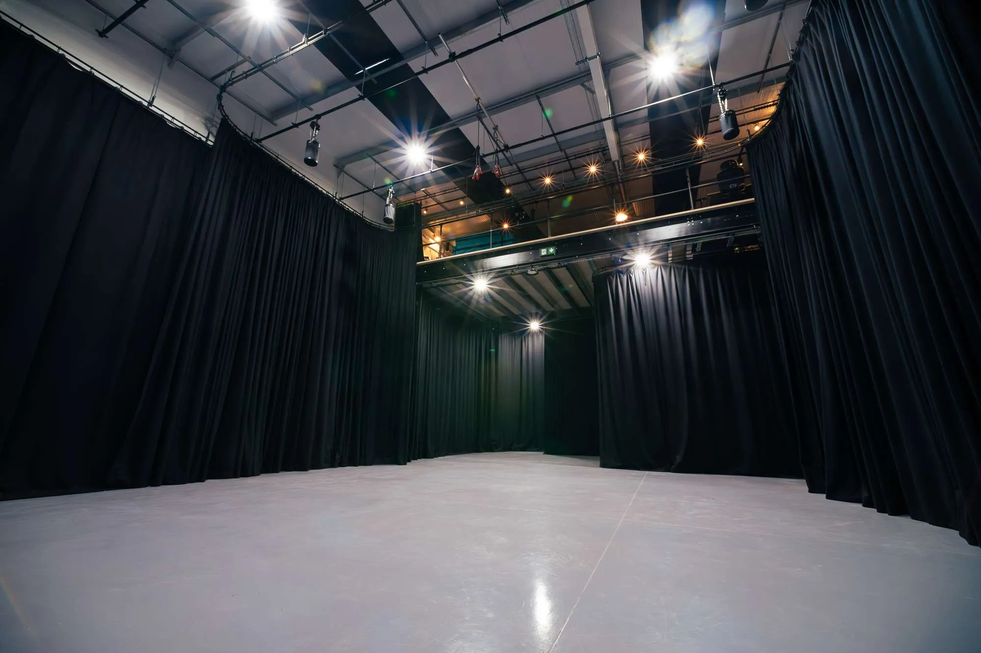 A spacious, empty film TV studio with a smooth, light gray floor, surrounded by black curtains and overhead stage lighting, giving the appearance of a photography or production space.