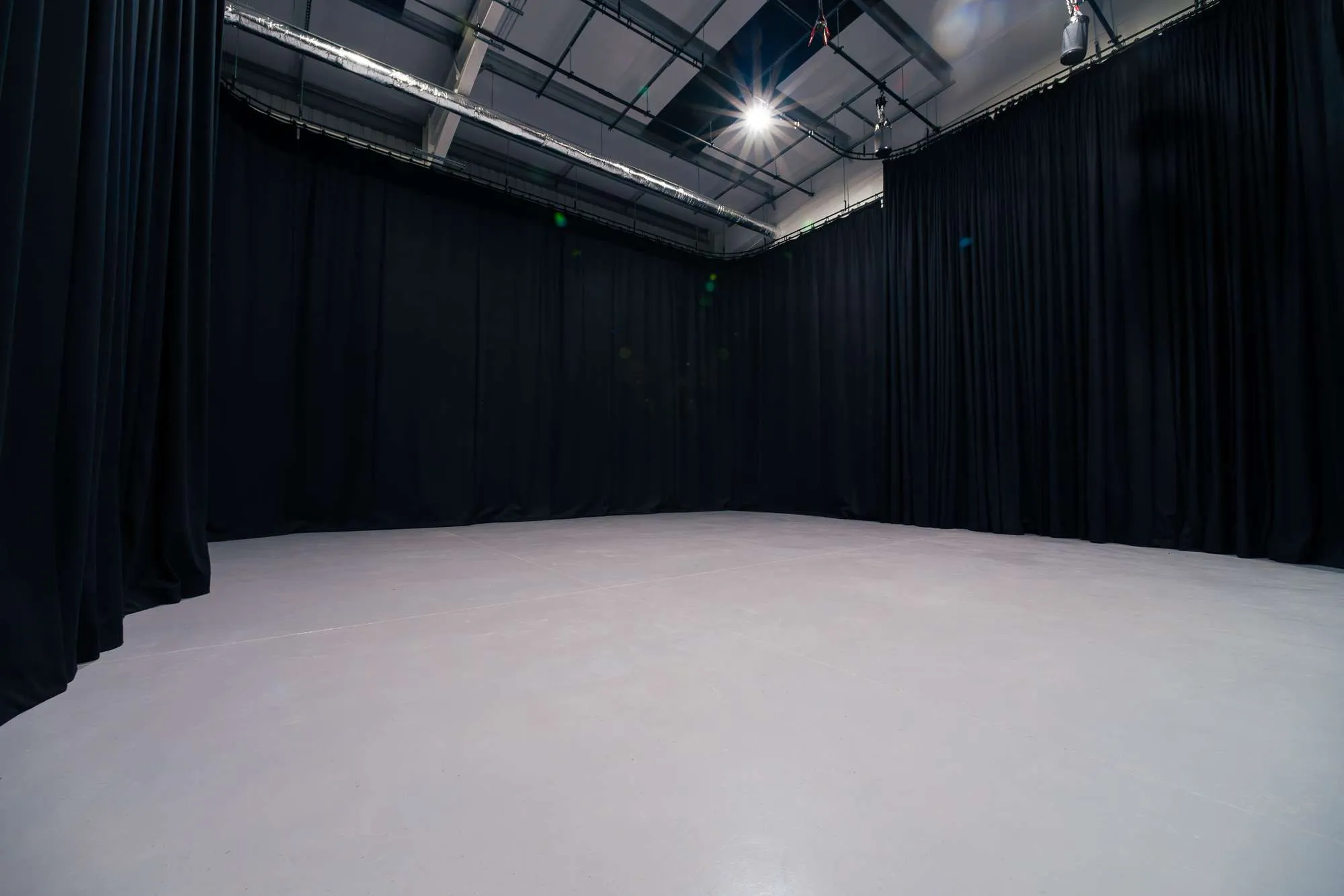 A spacious, empty film TV studio with a smooth, light gray floor, surrounded by black curtains and overhead stage lighting, giving the appearance of a photography or production space.